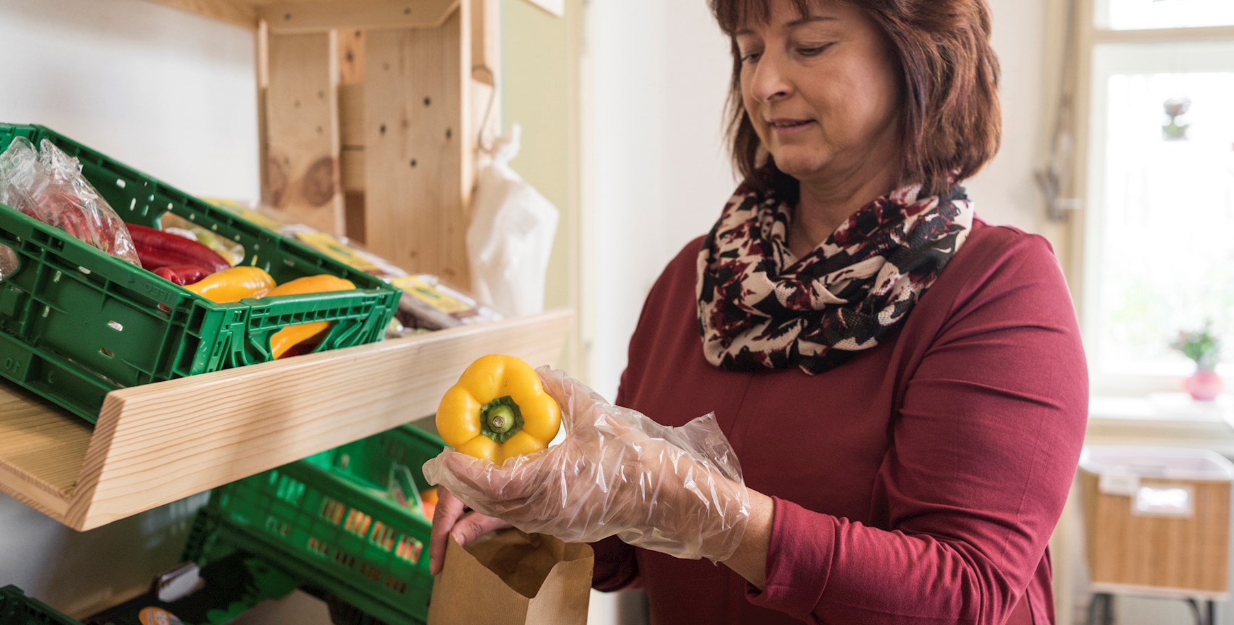 Eine Mitarbeiterin des Sozial­markts Fürstenfeld mit Einweg­handschuh gibt einen gelben Paprika in einen Sack aus Papier.