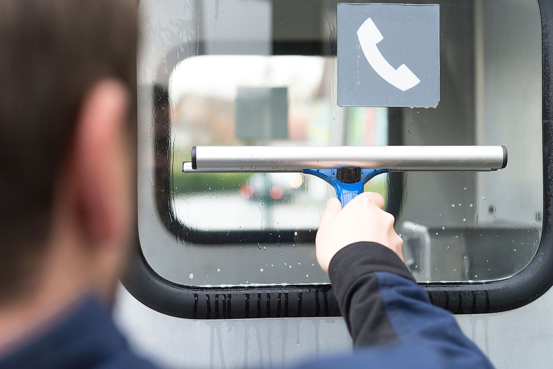 Mann führt einen Fenster-Abzieher über die Scheibe einer Telefonzelle.