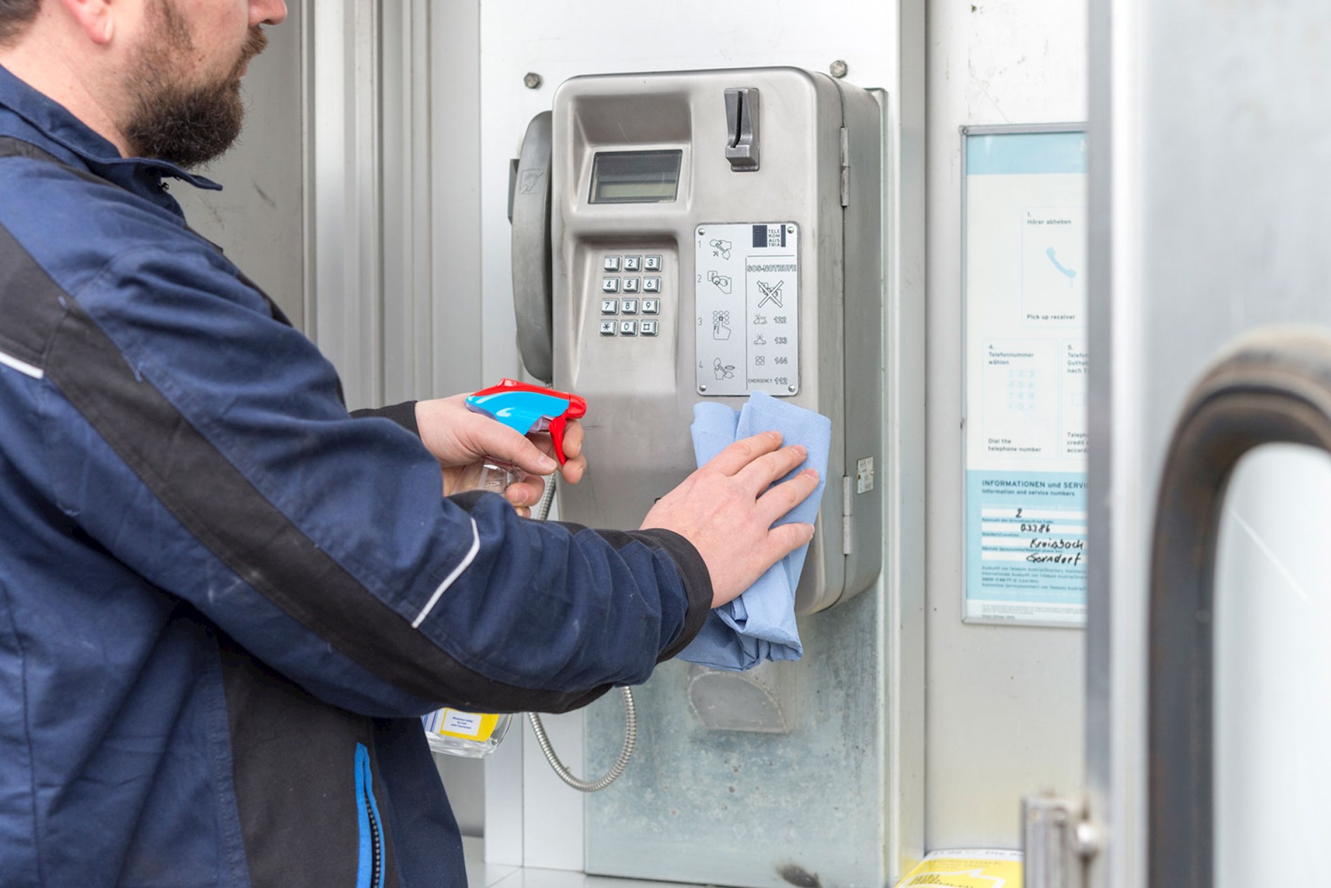 A person is cleaning the telephone in a phone booth. They are using cleaning products in a spray bottle and a light blue cloth.